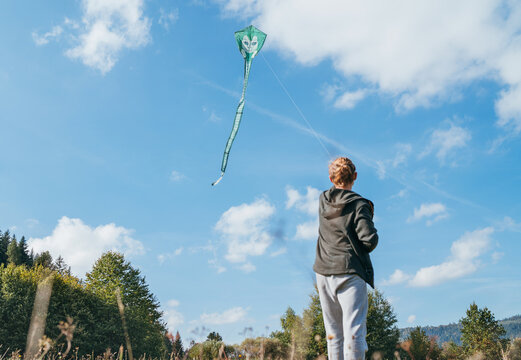 A young teenage boy with a flying green color kite toy on the high grass meadow in the mountain fields. Happy teenhood moments or outdoor time spending concept image.
