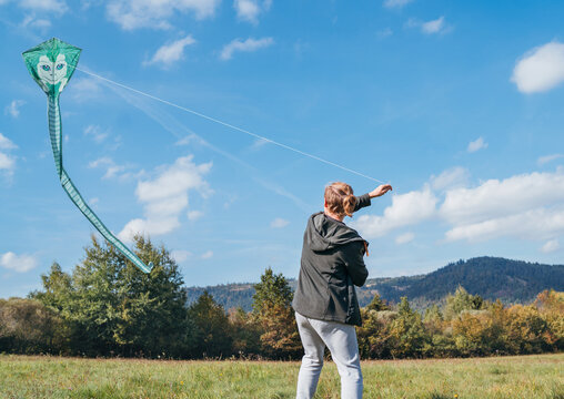 A young teenage boy with a flying green color kite toy on the high grass meadow in the mountain fields. Happy teenhood moments or outdoor time spending concept image.