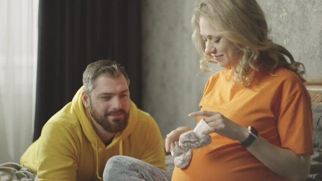 Happy pregnant couple laughing and having fun while waiting for the baby. The wife plays with booties, and the husband happily watches her. High quality 4k footage
