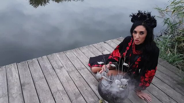A girl in an embroidered black and red dress with beads, in a black wreath, cooks with herbs in a cast-iron cauldron and conjures, sitting on a pier on the river bank against the background of reeds