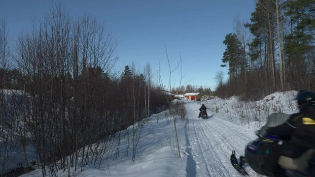 Two snowmobiles driving on a path with trees on both side, on cold sunny winter day in Sweden. Static shot.