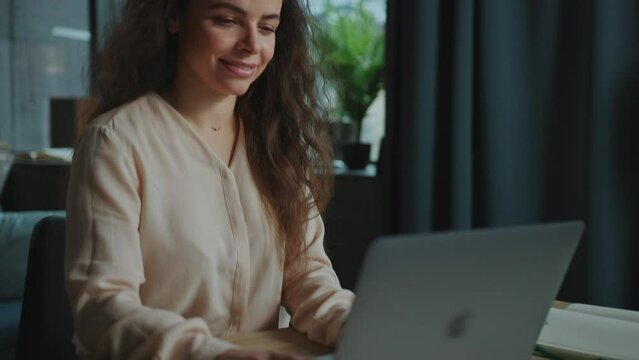 Close-up footage of woman's hands typing on keyboard of laptop. Attractive Caucasian woman with long curly hair using laptop, chatting, surfing social media feed and smiling indoors. Lifestyle concept