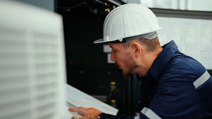 Caucasian production engineer in safety wear is reading the manual of a machine to find an error. A male factory worker is checking the industrial control panel of a robotic machine for maintenance.