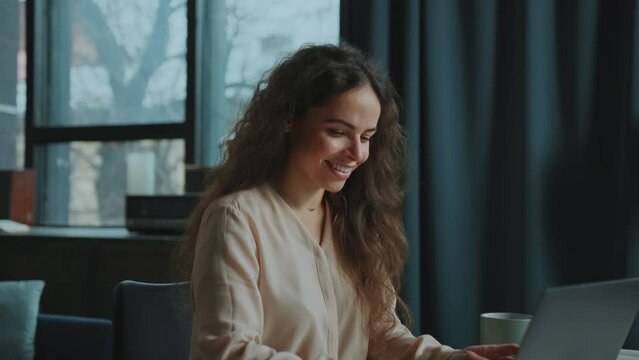Shot of joyful Caucasian young woman sitting at workplace with laptop, typing on keyboard in loft office. Portrait of curly girl smiling at camera. Indoors. Cozy interior