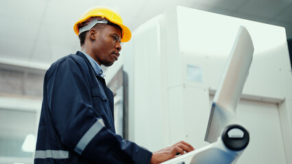 African production engineer in safety wear is operating CNC machine in the factory. A male factory worker with responsibility for programming industrial machine to control the process of production.