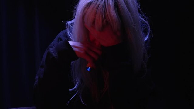 Depressed blonde woman hiding face stressfully smoking disposable vape while sitting in a dark room