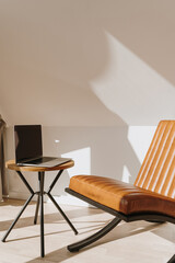 Aesthetic luxury modern home office workspace interior with laptop computer, orange leather armchair and coffee table. Sunlight shadows on the wall. Lounge space for relaxation and chill