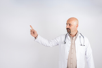 Showing copy space, portrait of smiling doctor showing copy space. Caucasian handsome male doctor in white coat with stethoscope pointing empty space with his finger. Isolated light gray background.