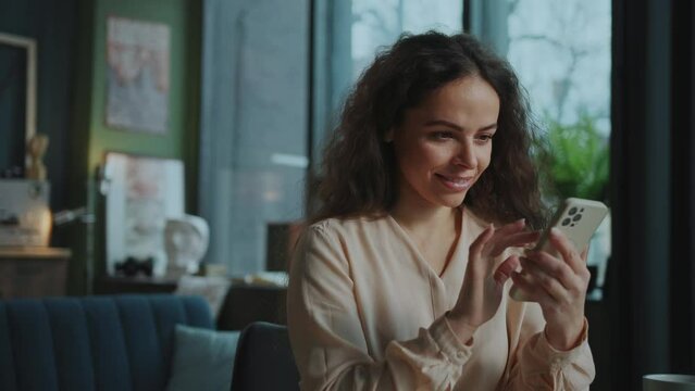Young smiling bloger woman enjoying surfing internet and posting on social media. Footage of joyful Caucasian girl with long natural curly hair looking at screen of smartphone. Online communication