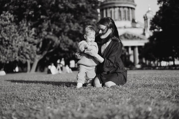 Caucasian mother of little son sitting on grass in front of st isaac cathedral helping boy stand...