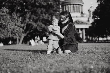 Caucasian mother of little son sitting on grass in front of st isaac cathedral helping boy stand holding by hand.
