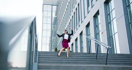 Caucasian pretty young business woman walking down the stairs on street in city dancing on headphones. Working financial manager.