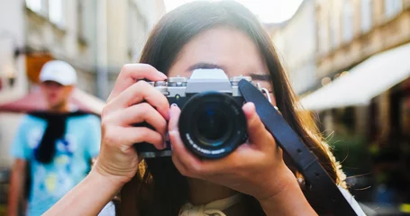 Fototapeten Wohnzimmer Close up portrait of cheerful beautiful female in good mood with camera standing outdoors in city. Happy pretty young Asian woman tourist taking photo. Photographer taking pictures. Urban concept  © ihorvsn