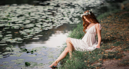 Beautiful red haired girl in white vintage dress and wreath of flowers sitting on the shore of river. Fairytale story. Warm art work.