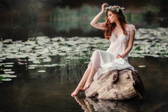 Beautiful red haired girl in white vintage dress and wreath of flowers sitting on the shore of river. Fairytale story. Warm art work.