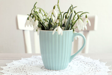  Spring card: a bouquet of snowdrops in a pastel mug against the background of a white chair, a white openwork napkin, space for text