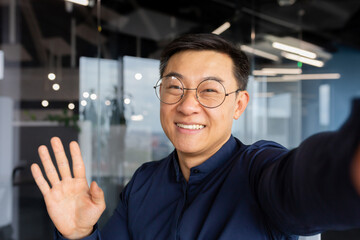 Asian man talking on video call using online communication app on phone, businessman in office taking selfie and smiling at camera, man greeting waving hand while talking with friends.