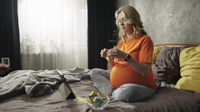 A pregnant woman looks at a tablet and eats a candy. The wife chooses a movie on the tablet and happily watches it. High quality 4k footage