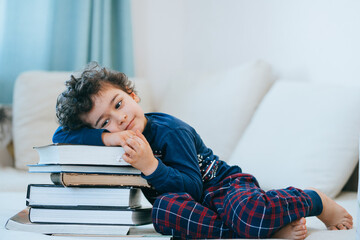Thoughtful little boy in pyjamas laying on couch leans on stack of books against copy space on background. Curly caucasian kid dreaming, smart toddler loves literature. Education, knowledge, childhood