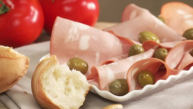 mortadella on a white plate with olives piece of bread and tomatoes. italian sausage on a wooden table with a napkin. serving meat delicacy. traditional boiled sausage under sunlight. rustic food.