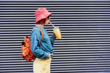 Stylish girl in bright clothes and bucket hat drinking sugar flavored tapioca bubble tea while...