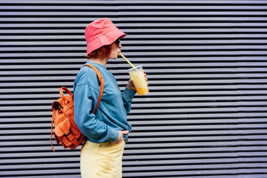Stylish girl in bright clothes and bucket hat drinking sugar flavored tapioca bubble tea while walking near gray striped urban wall. Portrait of fashionable hipster girl. Street fashion. Copy space.
