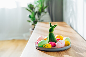 Easter home decor. Green bunny rabbit figurine and colored easter chocolate eggs on plate on wooden...