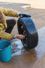 Outdoors, a man in a yellow dirty jacket with a hand without a glove washes a wheel from a car with a rag and water from a bucket, rinses the cracks, drops drip onto the mat, copy space.