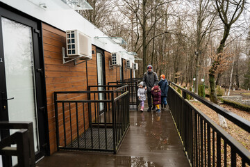 Father with three kids on terrace of one-storey modular houses in spring rainy forest.