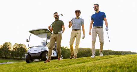 Three handsome caucasian men strolling with their clubs on the golf field near white golf car and talking. Outdoor