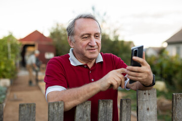 Mature man standing relaxed near fence talking on phone in backyard smiling