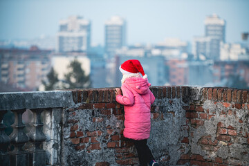  A little girl in a pink jacket and a New Year's cap looks out over the city in a light haze from above