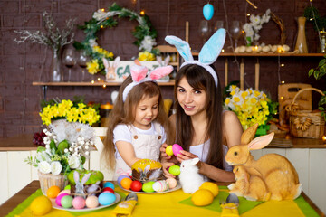 Fototapeta premium Easter, mom and daughter with Easter eggs and cake at the festive table are smiling and preparing for Easter at home in the kitchen in the ears of a hare.