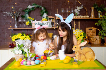 Easter, mom and daughter with Easter eggs and cake at the festive table are smiling and preparing for Easter at home in the kitchen in the ears of a hare.