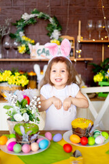 Obraz na płótnie Canvas easter, a little baby girl folded her hands like a bunny in the ears of a rabbit and smiles at home at a festive table with Easter eggs and a cake at home in the kitchen