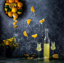 Limoncello in bottle and two grappas wineglass, fresh lemon in plate. Artistic still life in two places with fly and splash ingredients on dark background.