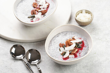 Breakfast idea - two bowls with with chia seeds pudding, red raspberry smoothie, pecan nuts, coconut flakes and pepitas on gray background