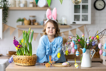 Portrait of cute pretty smiling little girl in blue dress and bunny rabbit ears preparing easter table decor. Fresh spring bright colourful flowers in the kitchen. Sunny day, family time