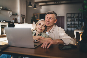 Happy aged man embracing woman while sitting at coffee table with laptop