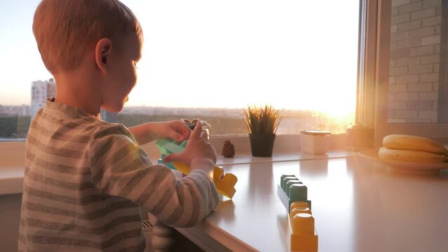 Little intelligent child plays children constructor at table by window at sunset shine. Inventive preschoolers funny boy collects crafts from details. Kids educational games. Dream of happy childhood