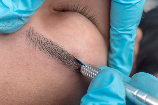 An esthetician using a microblade tool to make small cuts into the eyebrow of a customer. A Microblading technique to implant pigment.