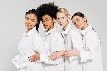 Obraz premium positive and sensual multicultural women looking at camera while posing in white shirts isolated on grey.