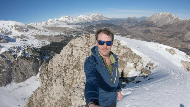 young caucasian man with sunglass and messy hairs man at the summit of a mountain. In background moutins peaks some snowy some not. Barges mountain in Devoluy. Selfie panoramic view in slow motion