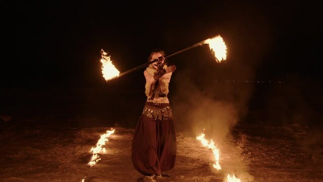 Mystical spectacular fire performance in night. Female artist in tribal costume goes along fiery path, twirls rod lit at ends in hands. Young attractive woman revolves flamy props outdoors