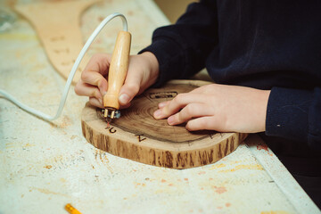 Boy burn out numbers with soldering iron on wooden disc. Kid makes wooden clock in the workshop.