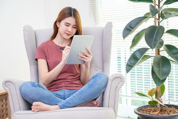 Asian woman sitting on yellow sofa using digital tablet and pen for learning shopping or working online near window