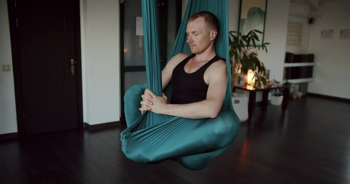 Man meditating in aerial hammock. Zoom out view of calm male sitting in green hammock and meditating with closed eyes during aerial yoga session in studio in morning.