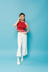 full length of positive asian woman posing with hand in pocket of white pants and talking on smartphone on blue background.