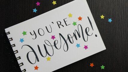 Fototapeta na wymiar YOU'RE AWESOME! lettering in notebook with colorful stars on black wooden desk