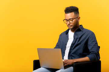 Concentrated indian guy using laptop in casual shirt and glasses using laptop sitting isolated on yellow background, freelancer guy is working remotely, developer coding, student studying on distance